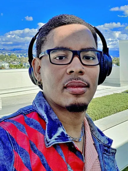 Manuel A. Dominguez in glasses and headphones, wearing a red and blue jacket, on a rooftop under a blue sky.